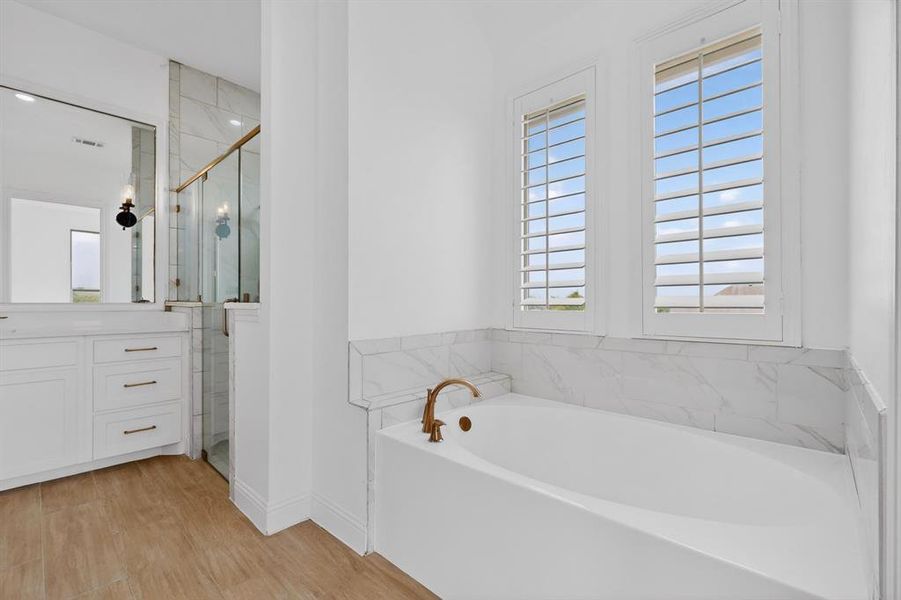 Soaking tub with brass-finish fixtures, dual windows with plantation shutters, tile surround, wood-finish flooring, and a vanity with a quartz countertop Soaking tub with brass-finish fixtures, dual windows with plantation shutters, tile surround, wood-finish flooring, and a vanity with a quartz countertop