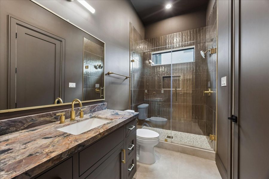 Rich stone surfaces, dramatic full height tile, and warm brass fixtures give this ensuite bath a distinctive designer feel. A frameless glass shower with built in niche and transom window completes the sophisticated space.