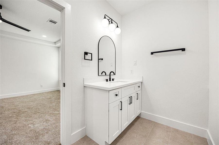 Primary Bedroom-This photo shows a modern bathroom with a white vanity featuring a black faucet and hardware. An oval mirror and dual lights adorn the wall above the sink. The adjacent room is carpeted, suggesting a bedroom or living space. The overall look is clean and contemporary.