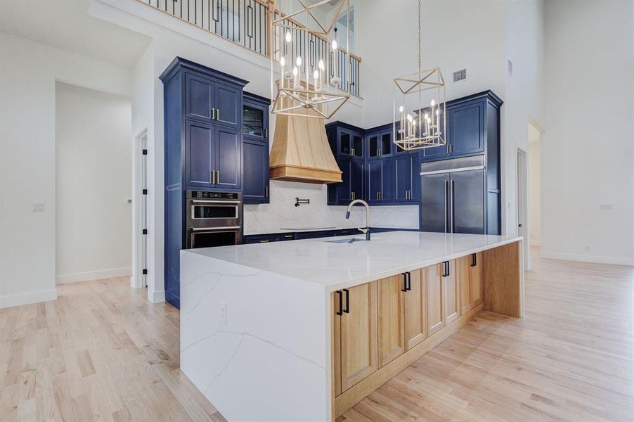 Spacious kitchen featuring a large central island with a waterfall countertop, two-tone cabinetry in blue and natural wood, stainless steel built-in appliances, light wood-finish flooring, and high ceilings with decorative light fixtures
