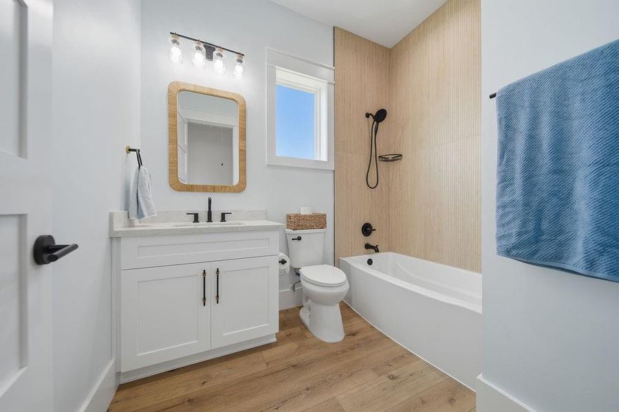 Bright, modern bathroom with a sleek white vanity, wood-accented mirror, and contemporary fixtures. Features a bathtub with a stylish wood-look tiled wall and a large window for natural light. The space is enhanced by light wood flooring and a tidy, minimalist design.