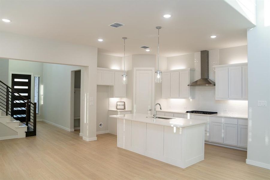 Open-concept kitchen featuring an island with an integrated sink, white cabinetry, a stainless steel range hood, and a gas cooktop Open-concept kitchen featuring an island with an integrated sink, white cabinetry, a stainless steel range hood, and a gas cooktop