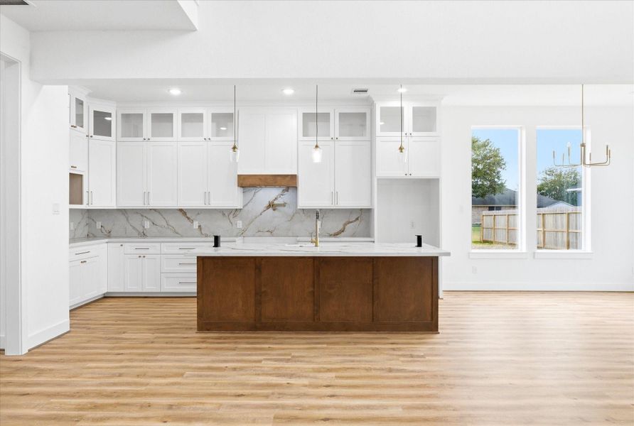 A brightly lit kitchen showcasing a large quartz island, solid wood cabinets with integrated soft-close hinges are visible beneath a large, decorative vent hood. A stainless steel pot filler is mounted over a 36 inch gas range.