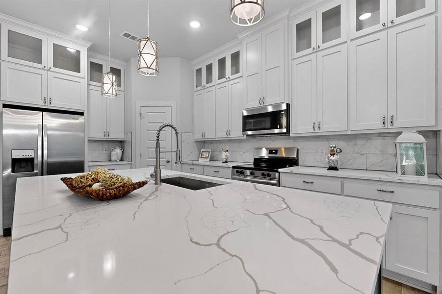 Kitchen featuring light stone counters, decorative backsplash, hanging light fixtures, stainless steel appliances, and glass insert cabinets Kitchen featuring light stone counters, decorative backsplash, hanging light fixtures, stainless steel appliances, and glass insert cabinets