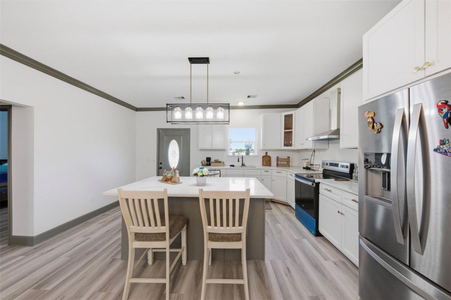 Modern kitchen with sleek white cabinetry, stainless steel appliances, and a central island with seating. The space is well-lit with a contemporary light fixture and features light wood flooring, creating a bright and inviting atmosphere.