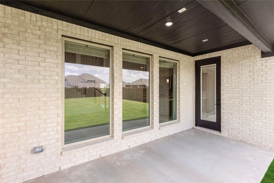 Covered back patio with light brick exterior, large windows, dark entry door, and concrete floor overlooking grassy backyard.