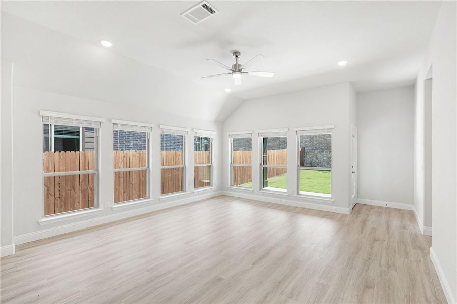 Empty living room with vaulted ceiling, ceiling fan, light wood floors, and a row of windows overlooking a backyard fence. Empty living room with vaulted ceiling, ceiling fan, light wood floors, and a row of windows overlooking a backyard fence.