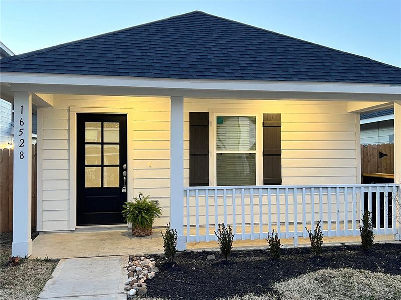 This is a charming, single-story home featuring a clean, white exterior with a contrasting black front door and dark roof shingles. It includes a small front porch with a railing, accented by modest landscaping and a well-maintained lawn.