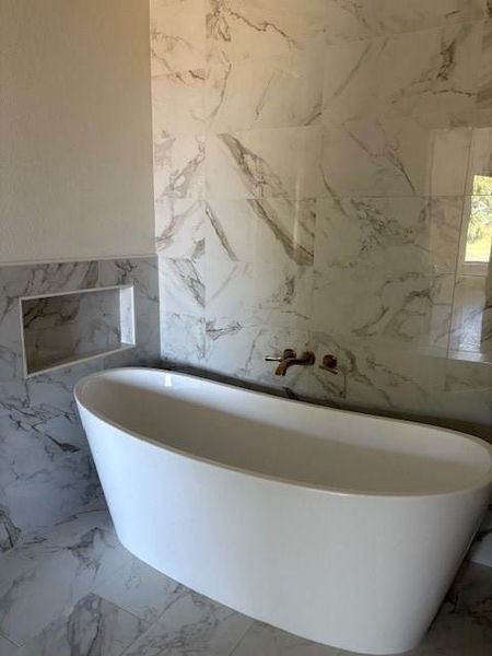 Full bath with light marble finish flooring, a freestanding bath, and tile walls