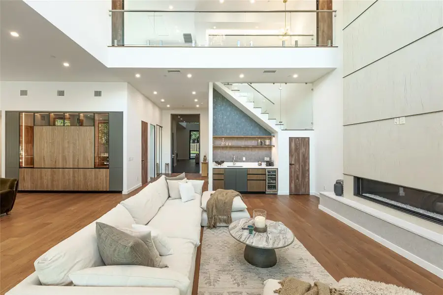 A spacious living room features soaring ceilings, a modern fireplace, and rich hardwood flooring leading to an elegant wet bar and staircase.