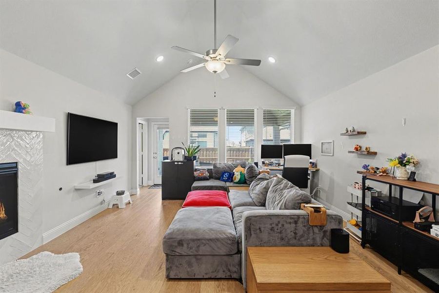 Spacious living area featuring a vaulted ceiling with a ceiling fan, light wood-finish flooring, a contemporary fireplace with a herringbone tile surround and white mantel, and multiple windows allowing natural light