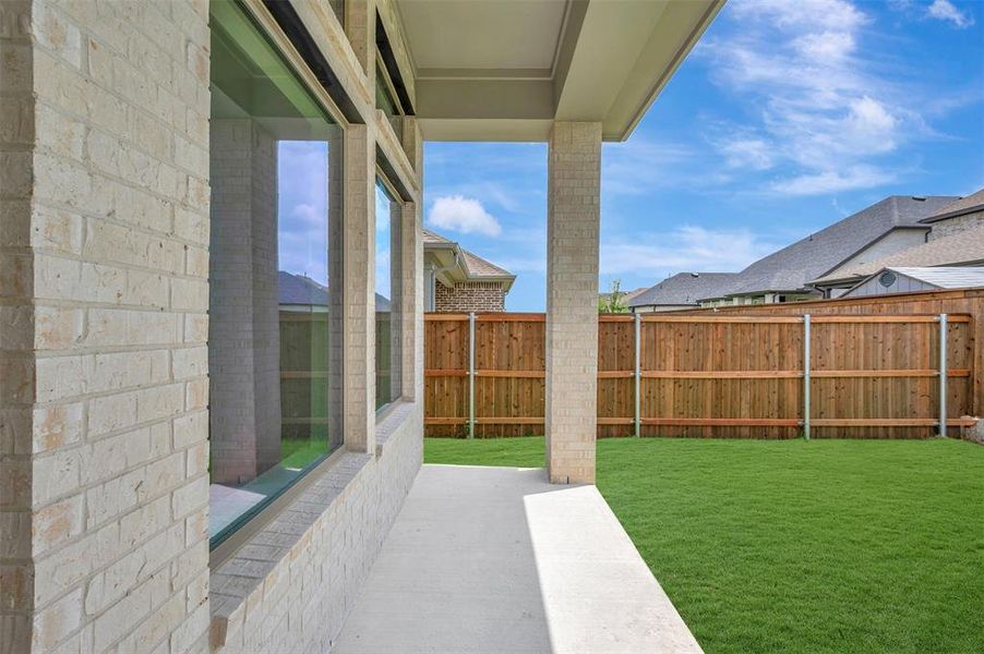 Covered patio with light-toned brick, full-height windows, a concrete walkway, and a private wooden fence enclosing a grass lawn