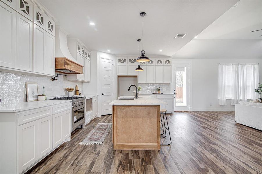 Kitchen with custom exhaust hood, a kitchen island with sink, high end stainless steel range oven, decorative light fixtures, and white cabinetry