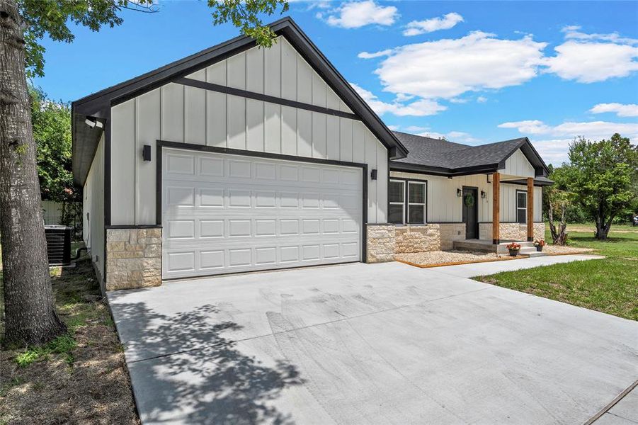 Modern farmhouse exterior with white vertical siding, black trim accents, a two-car garage, stone veneer skirting, and a concrete driveway Modern farmhouse exterior with white vertical siding, black trim accents, a two-car garage, stone veneer skirting, and a concrete driveway