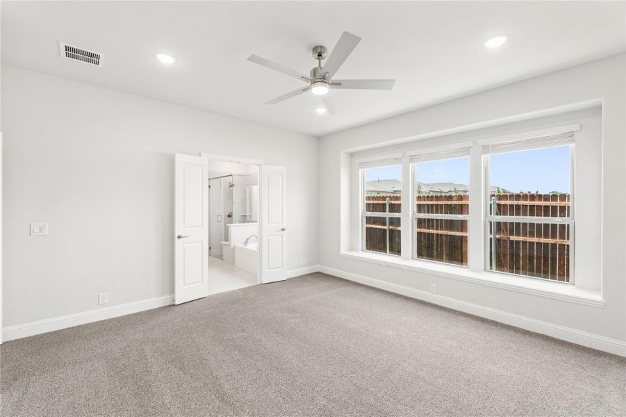 Empty bedroom with carpet, ceiling fan, large windows, and ensuite bathroom door open