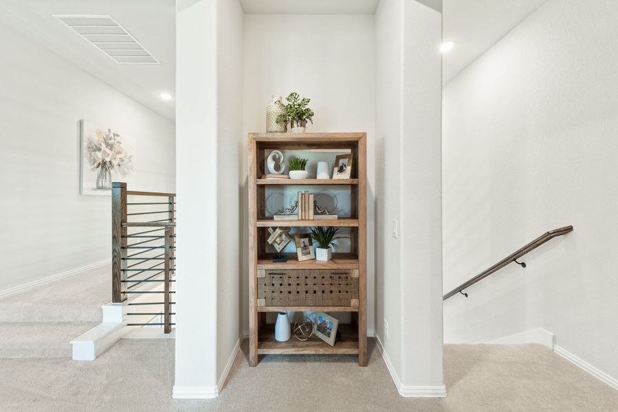 Upstairs hallway landing with wooden bookshelf displaying decor and staircase with metal railing in background