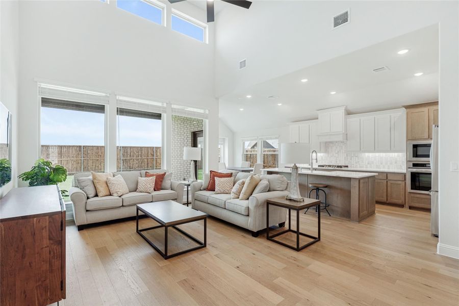 Open-concept living room with two sofas and high ceilings, flowing into kitchen with white cabinets and island, hardwood floors throughout