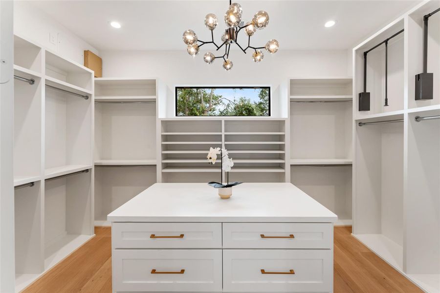 The larger of two walk-in closets in the primary suite functions as a true dressing room, complete with extensive storage, center island workspace, natural light, and elegant chandelier lighting.