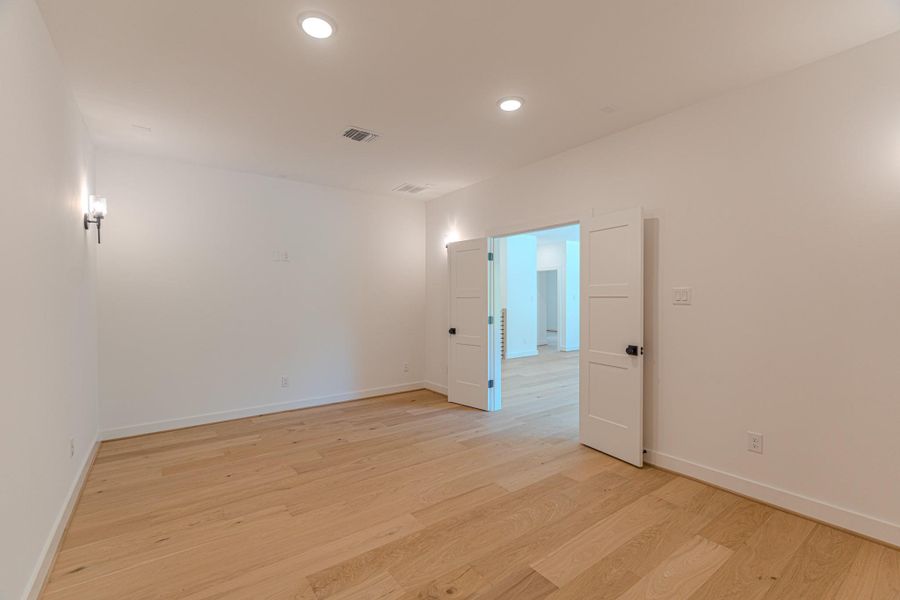 This photo showcases a spacious, well-lit room with light wood flooring and white walls. It features recessed lighting and wall sconces, with double doors leading to an adjacent area, suggesting an open and inviting layout.