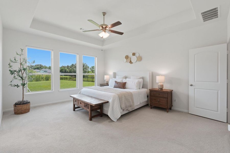 This spacious, bright bedroom features large windows with serene outdoor views, a ceiling fan, and neutral decor. It includes a cozy bed with side tables and lamps, perfect for a relaxing retreat.