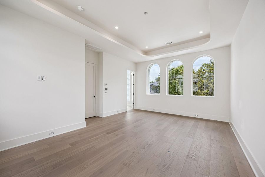 Spacious Primary Bedroom with a multi-tiered tray ceiling with gently rounded corners. A trio of vertically proportioned arched windows introduces a classical influence on both north and south side of the room. Construction as of June 1st, 2026.