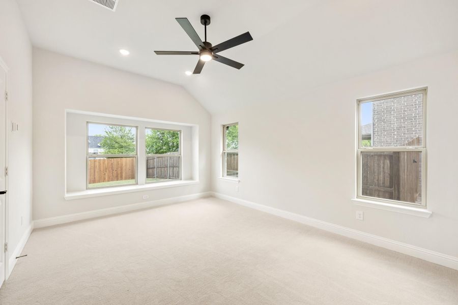 Empty bedroom with vaulted ceiling, ceiling fan, carpet flooring, and multiple windows with backyard views