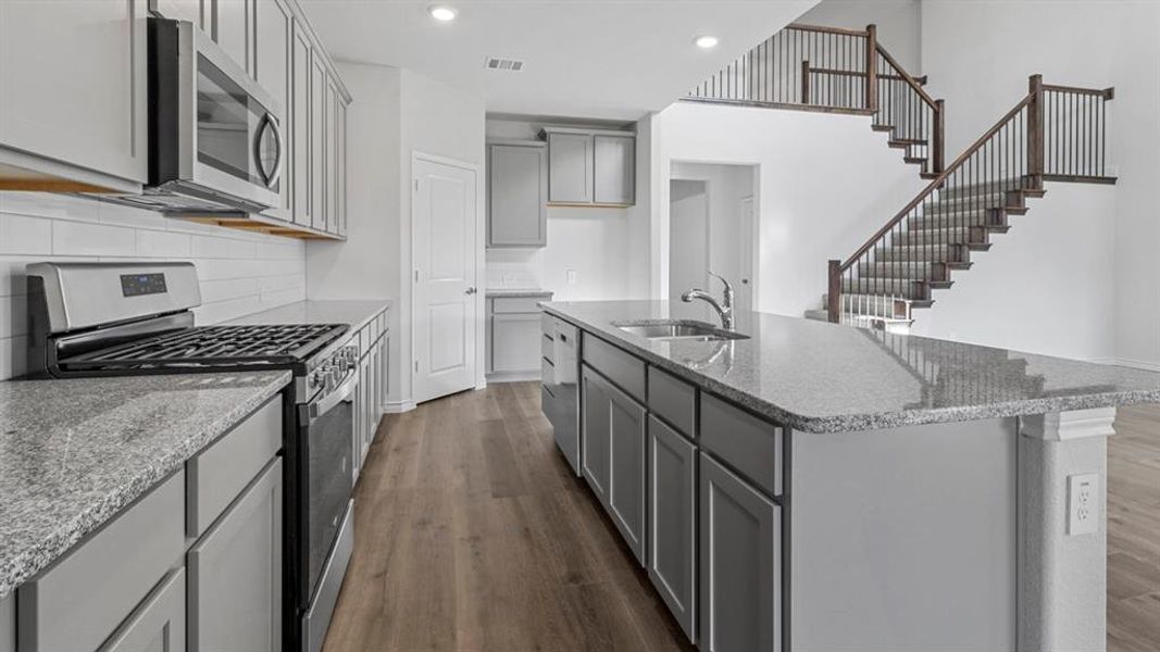 Kitchen featuring gray cabinetry, appliances with stainless steel finishes, light stone countertops, an island with sink, and dark wood-style flooring