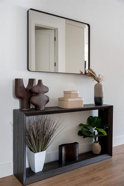 Rectangular wall-mounted mirror with a black frame above a wood-finish console table featuring two open shelves and a contrasting light wood-finish flooring