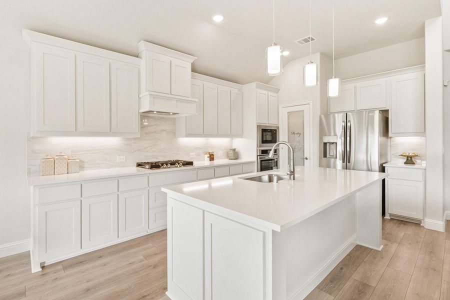 White kitchen with large island, quartz countertops, stainless steel appliances, and pendant lights over hardwood floors White kitchen with large island, quartz countertops, stainless steel appliances, and pendant lights over hardwood floors