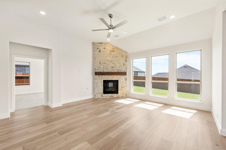 Open living room with stone fireplace, light hardwood floors, ceiling fan, and large windows Open living room with stone fireplace, light hardwood floors, ceiling fan, and large windows