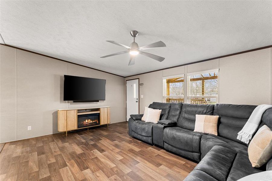 Living room with ornamental molding, wood finished floors, a ceiling fan, a textured ceiling, and a glass covered fireplace