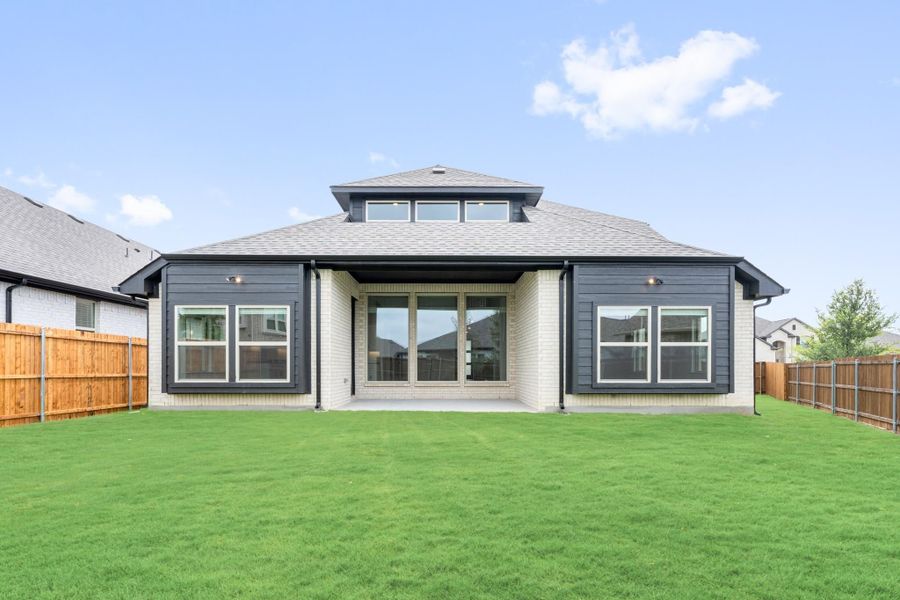 Rear exterior of single-story home with dark siding, brick accents, covered patio, and fenced backyard with green lawn. Rear exterior of single-story home with dark siding, brick accents, covered patio, and fenced backyard with green lawn.