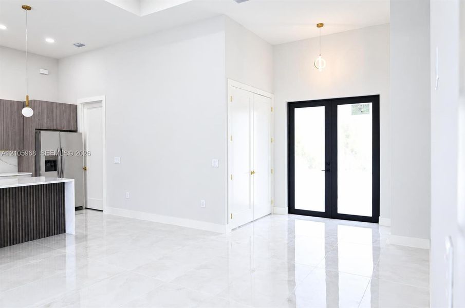 Open-concept interior featuring large-format polished tile flooring, high ceilings, a kitchen island with vertical wood-finish paneling, custom cabinetry, and black-framed glass-paneled double doors