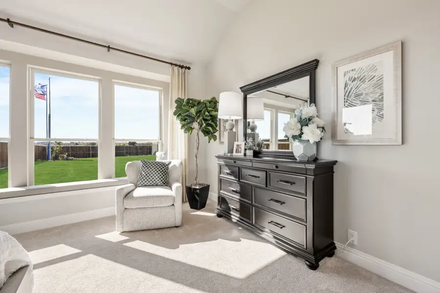 Bedroom with dark wood dresser and mirror, white armchair, vaulted ceiling, and large windows overlooking backyard