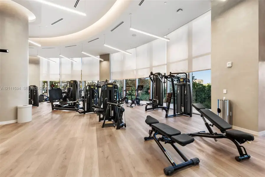 Fitness center featuring wood-finish flooring, floor-to-ceiling windows with roller shades, recessed cove lighting, and suspended linear LED fixtures Fitness center featuring wood-finish flooring, floor-to-ceiling windows with roller shades, recessed cove lighting, and suspended linear LED fixtures