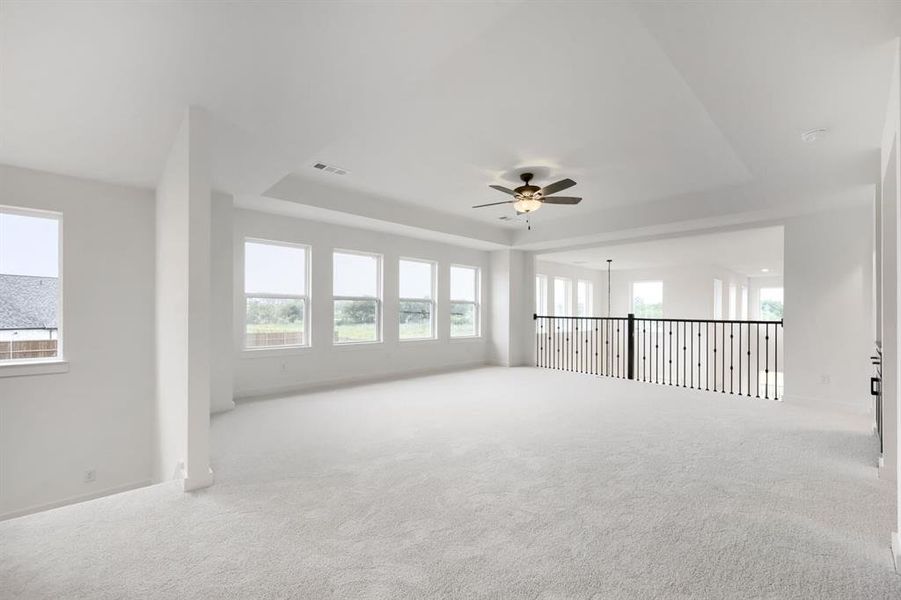 Spacious loft area featuring light carpet flooring, a multi-window wall, and a tray ceiling with a ceiling fan