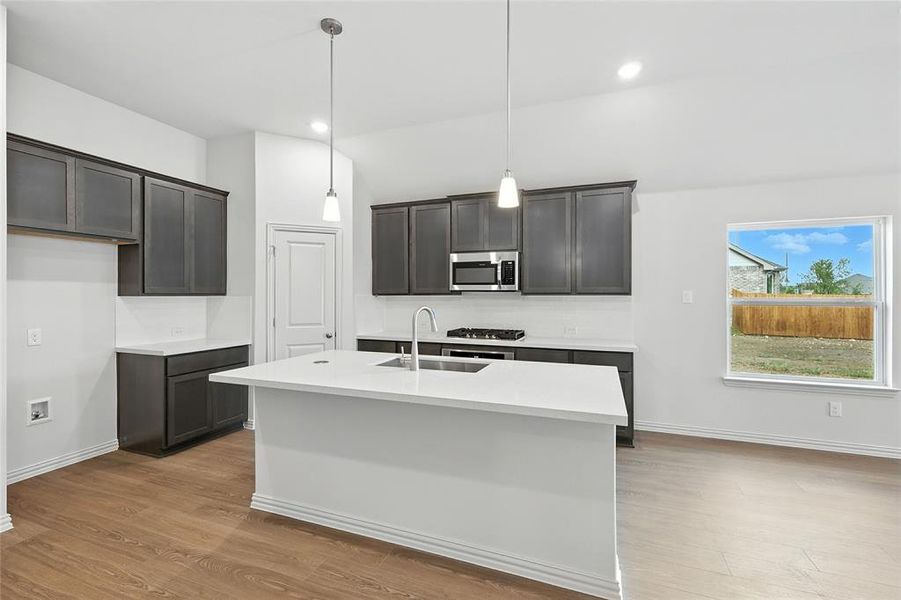 Kitchen featuring light wood-type flooring, pendant lighting, a kitchen island with sink, appliances with stainless steel finishes, and light stone counters Kitchen featuring light wood-type flooring, pendant lighting, a kitchen island with sink, appliances with stainless steel finishes, and light stone counters