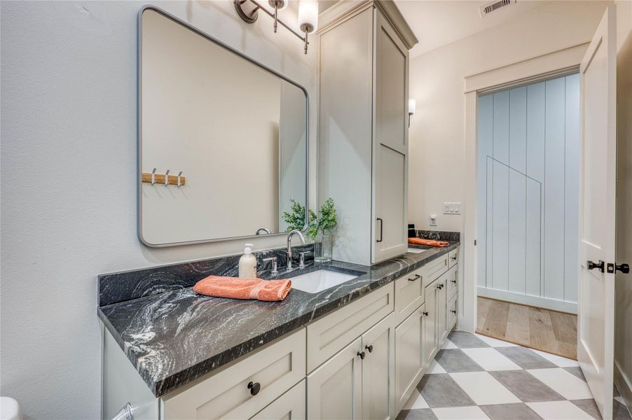 Bathroom featuring a gray and white checkered tile floor, light-colored vanity cabinetry with black hardware, a black patterned countertop, a built-in sink with a chrome faucet, and an upper built-in cabinet
