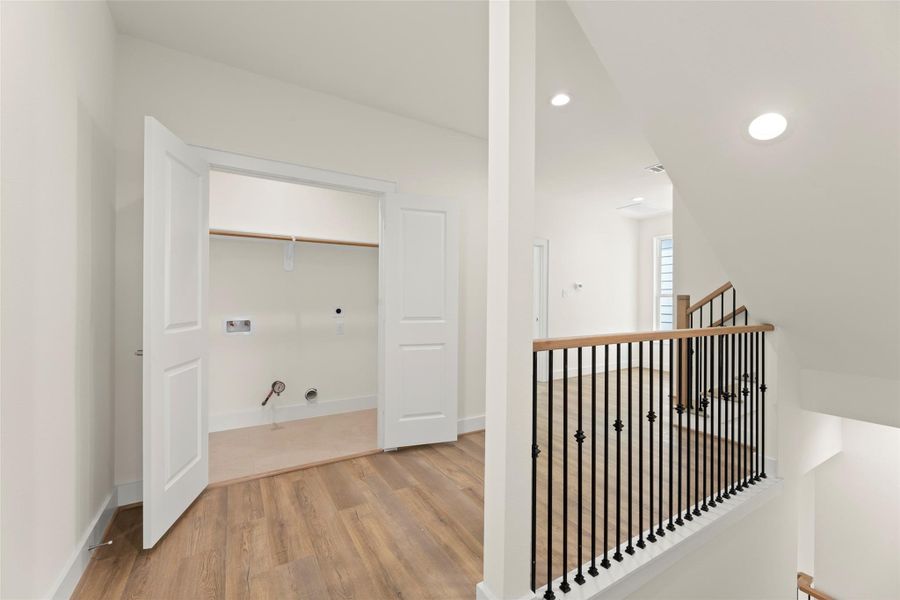 This photo features a bright, open landing area with wood flooring, leading to a stairway with elegant black railings. A utility closet is visible, ready for laundry appliances. The space is well-lit with recessed lighting, offering a modern and clean aesthetic.