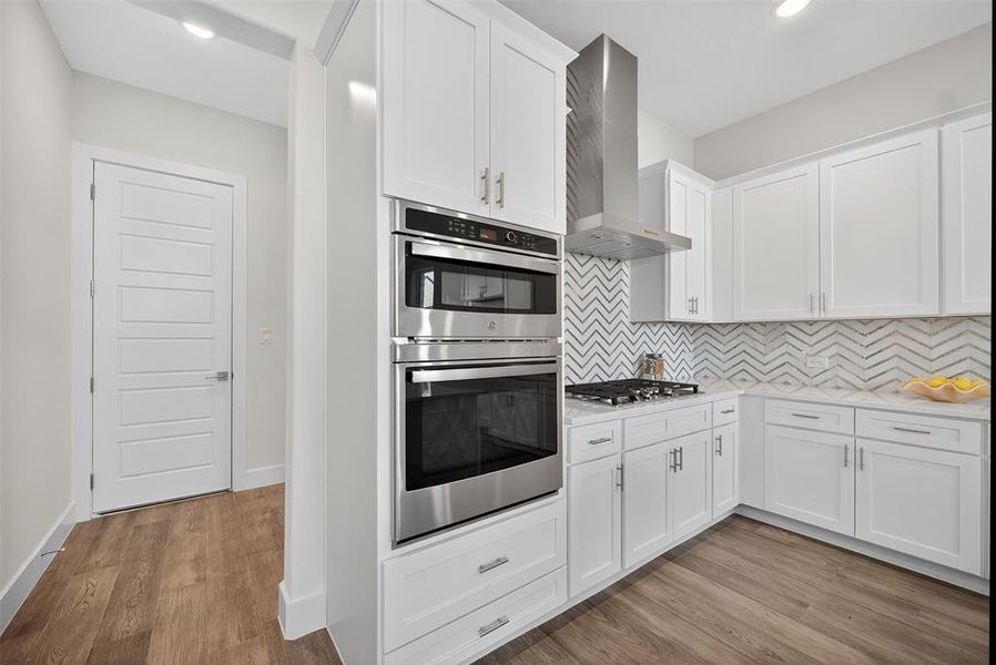 Kitchen featuring decorative backsplash, stainless steel appliances, white cabinets, wall chimney exhaust hood, and light wood finished floors Kitchen featuring decorative backsplash, stainless steel appliances, white cabinets, wall chimney exhaust hood, and light wood finished floors