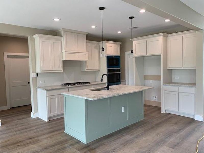 Kitchen featuring decorative light fixtures, light wood-type flooring, an island with sink, black appliances, and light stone countertops