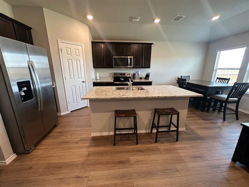 Kitchen featuring stainless steel appliances, dark wood-style floors, light stone counters, a breakfast bar, and an island with sink