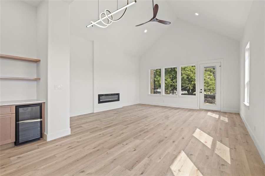 Unfurnished living room featuring high vaulted ceiling, beverage cooler, a glass covered fireplace, light wood-style floors, and ceiling fan