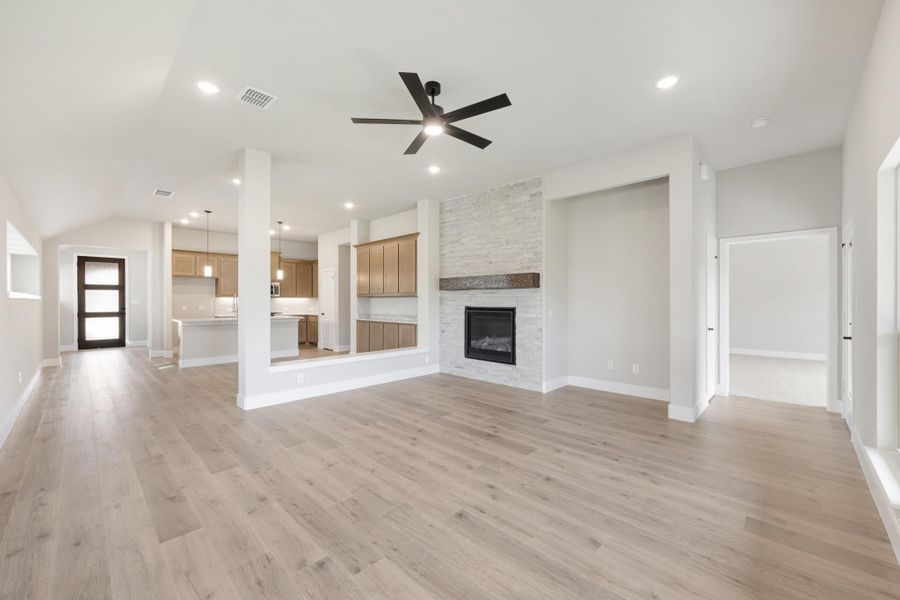 Open living area with stone fireplace, ceiling fan, hardwood floors, and open kitchen in background