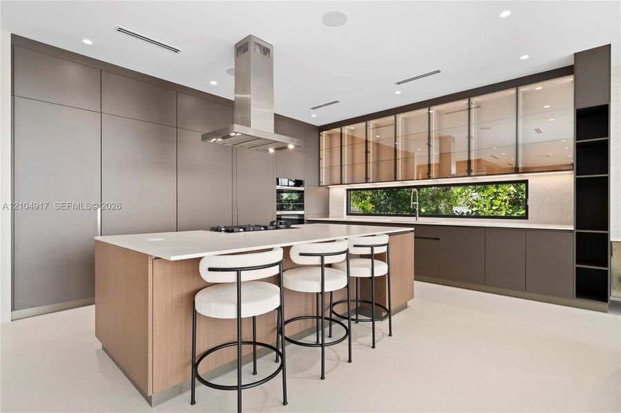 Contemporary kitchen with a large central island, stainless steel range hood, integrated appliances, and extensive cabinetry featuring glass-front uppers