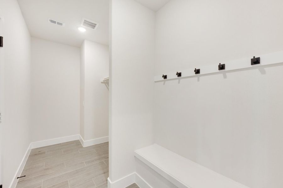 Mudroom with built-in bench, row of dark metal hooks, white walls, and light tile flooring