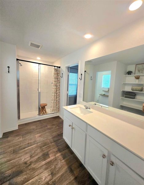 Bathroom featuring wood-finish flooring, a double vanity with white cabinetry and integrated sinks, a large mirror, and a glass-enclosed shower