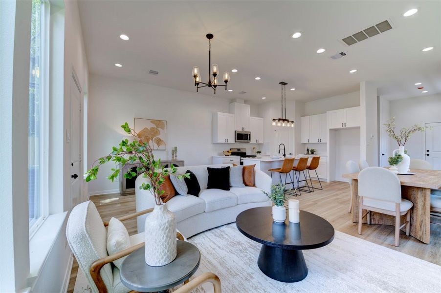 Open-concept first floor with hardwood floors, high ceilings, and recessed lighting throughout. Living area features a matte black chandelier and floor-to-ceiling windows. Dining table with seating for six sits to the right. Kitchen in the background shows white shaker cabinets, stainless appliances, island with leather bar stools, Edison rail pendant, and pantry niche. All three spaces flow seamlessly with no walls separating them.