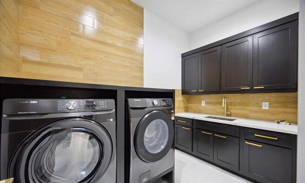 This is a modern laundry room featuring sleek dark cabinetry with gold hardware, a built-in sink, and high-end appliances. The bamboo backsplash adds a warm touch to the space.