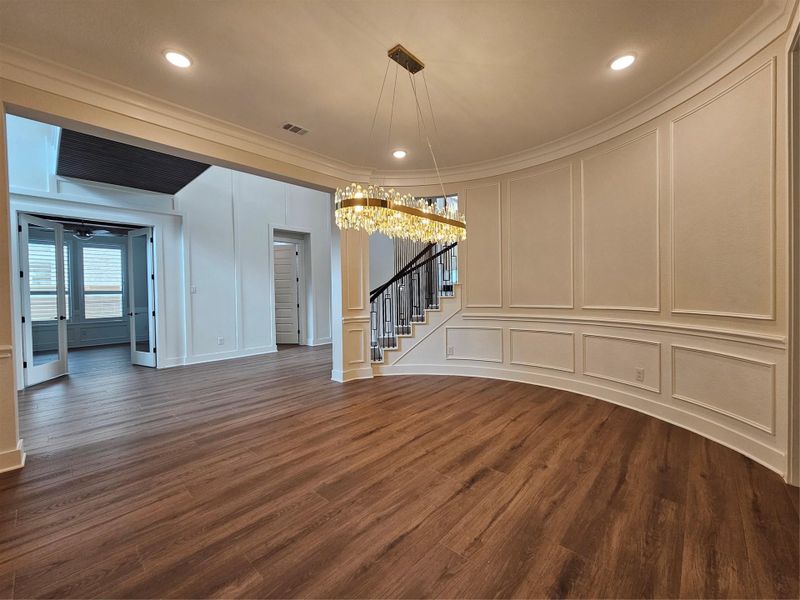 This photo features a spacious room with elegant wood flooring and detailed wall paneling. A striking chandelier hangs from the ceiling, illuminating the area. The space is open, leading to a staircase with black railings and a room with glass double doors, contributing to a modern and sophisticated atmosphere. This photo features a spacious room with elegant wood flooring and detailed wall paneling. A striking chandelier hangs from the ceiling, illuminating the area. The space is open, leading to a staircase with black railings and a room with glass double doors, contributing to a modern and sophisticated atmosphere.